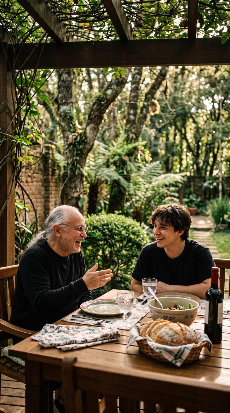 Pai e filho conversando e rindo à mesa de um almoço ao ar livre, cercados de verde